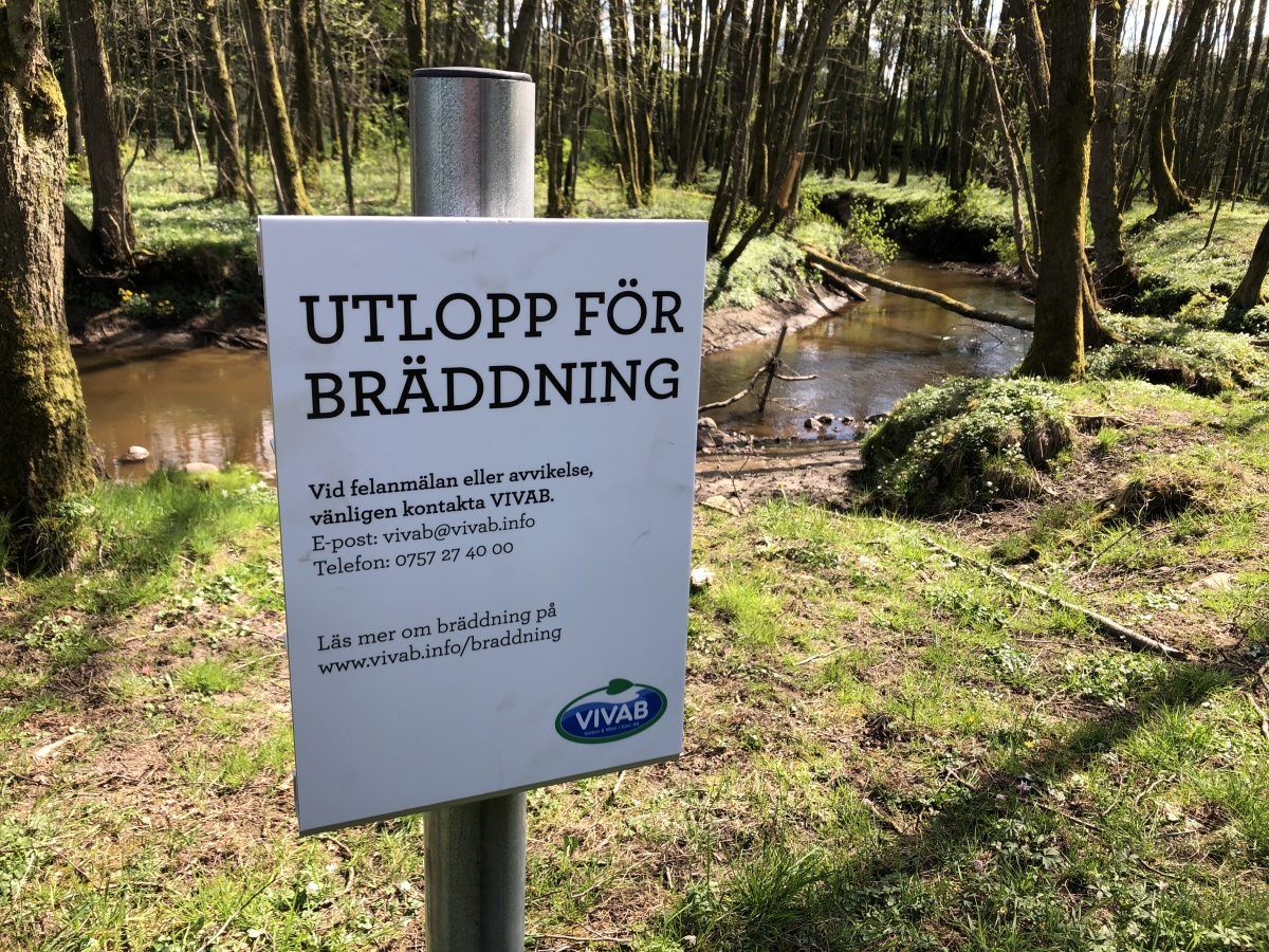 Skylt för utlopp för bräddning i förgrunden, en bäck i bakgrunden.